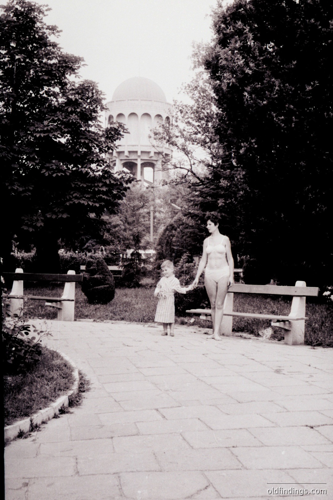 A woman and a young child walk hand-in-hand along a paved path, framed by lush greenery. A large, classical building with a dome is visible in the background. Likely a resort or public garden. The photograph’s style and subject suggest a 1960s or 70s vacation snapshot.