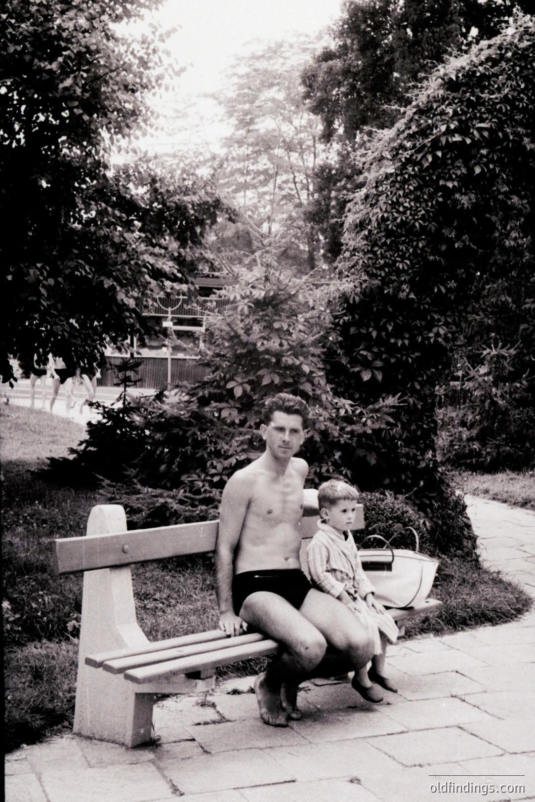 A man, bare-chested and wearing swim trunks, sits on a concrete bench with a young child dressed in a light-colored outfit. Lush foliage forms a backdrop. Likely a seaside resort setting, possibly Bulgaria. Photograph’s style suggests a candid moment, potentially from the 1960s or 70s.