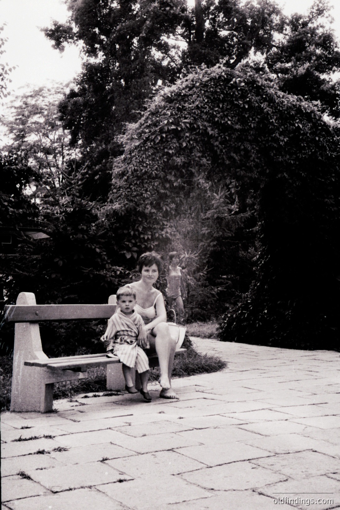 A mother and child sit on a stone bench, framed by dense foliage. The child wears a short patterned dress, and the woman is in a simple, sleeveless garment. The scene conveys a tranquil, domestic moment, likely 1960s or 70s. A paved path leads into the background.