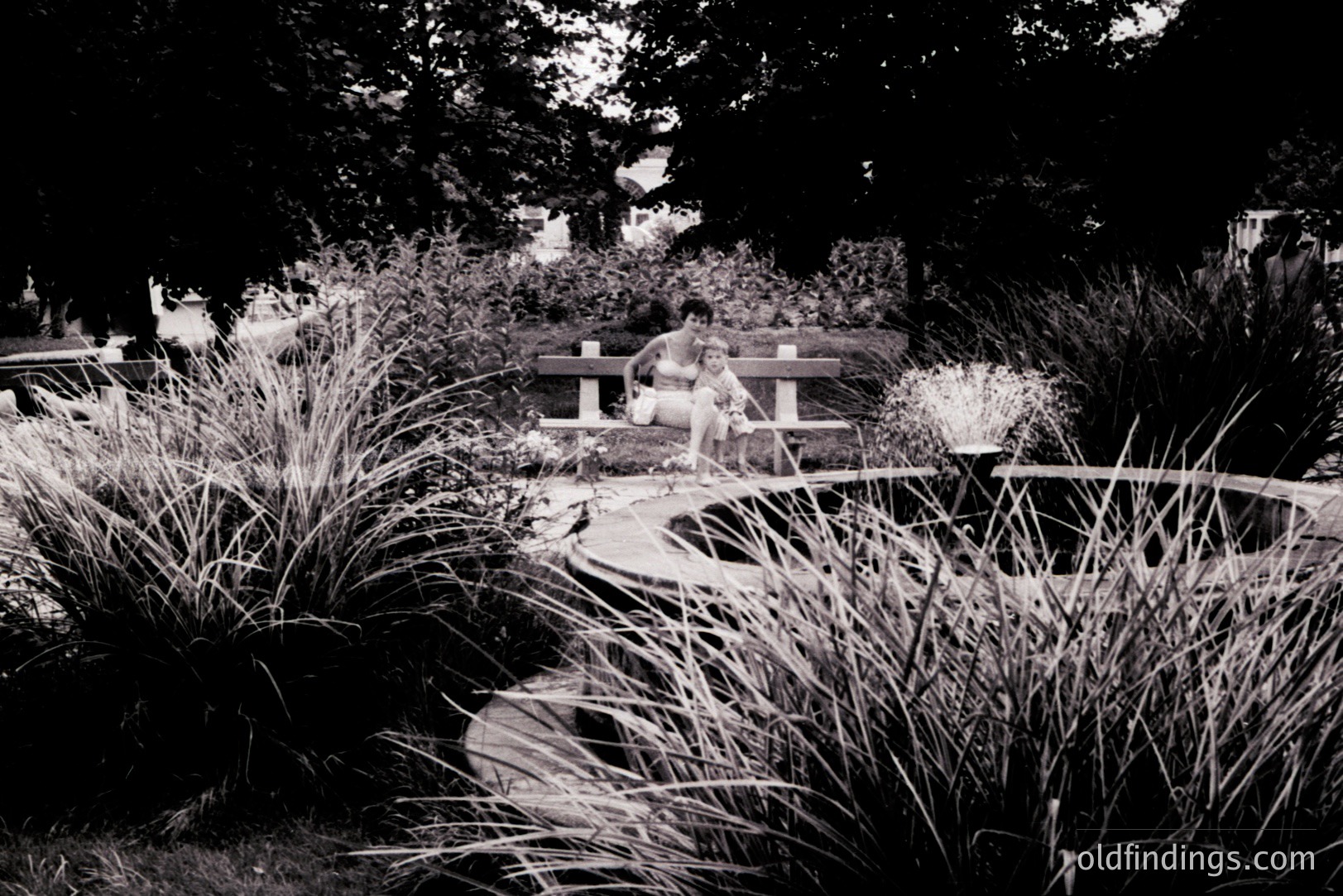 A woman and child seated on a bench overlook a circular fountain within a manicured garden. The architecture suggests a European park, likely 1970s-era, with formal plantings. Strong textural contrast between foliage and stonework. A serene, quiet moment captured in black and white. Stock potential for travel/lifestyle content.