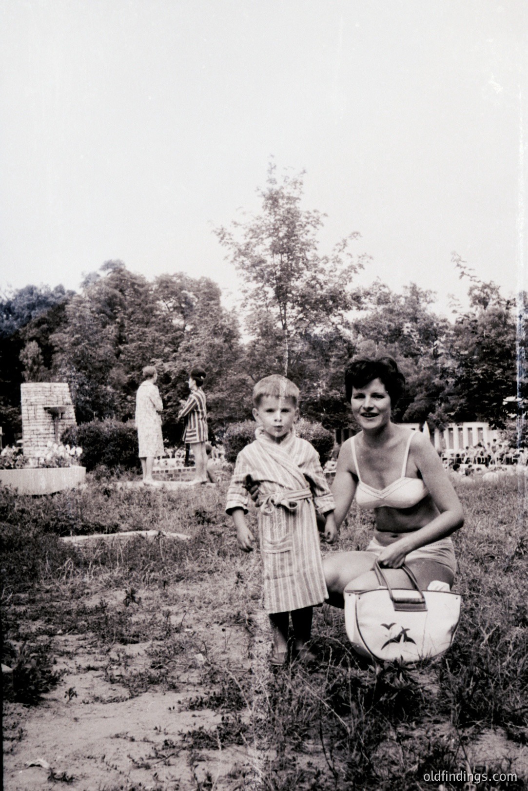 A young boy in a striped robe stands with a woman in a vintage bikini, seated near a travel bag. A classical ruin/statue is visible in the blurred background. Likely a vacation photo, possibly Bulgaria or a similar Mediterranean region, circa 1960s. Nostalgic snapshot.