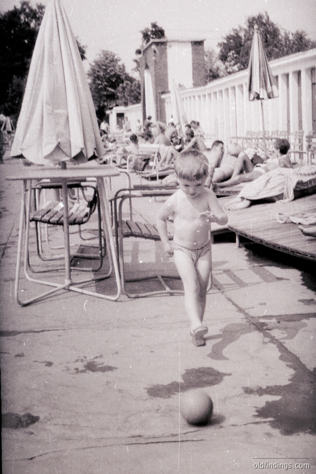 A young boy, scantily clad, runs on a wet concrete patio toward the camera, a ball at his feet. Behind him, a poolside scene with sunbathers and a simple architectural style suggests a resort or public pool. Likely 1960s, potentially Eastern European given the architecture. Evokes mid-century leisure and family vacations.