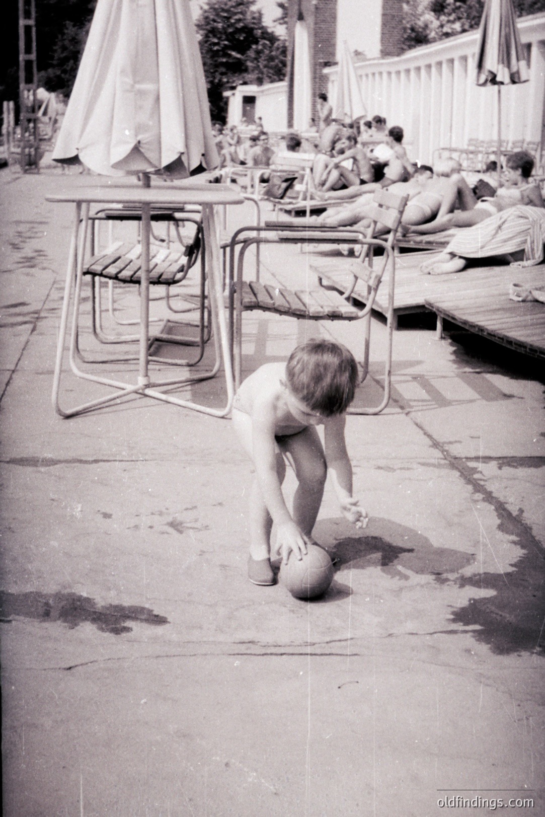 A young boy, partially obscured by patio furniture, crouches playfully near a ball on a concrete terrace. Behind him, figures lounge in swimwear; an outdoor pool and adjacent building suggest a resort setting. The photograph’s grainy quality and clothing styles suggest a mid-20th century (likely 1960s) snapshot. Likely an amateur holiday photo.