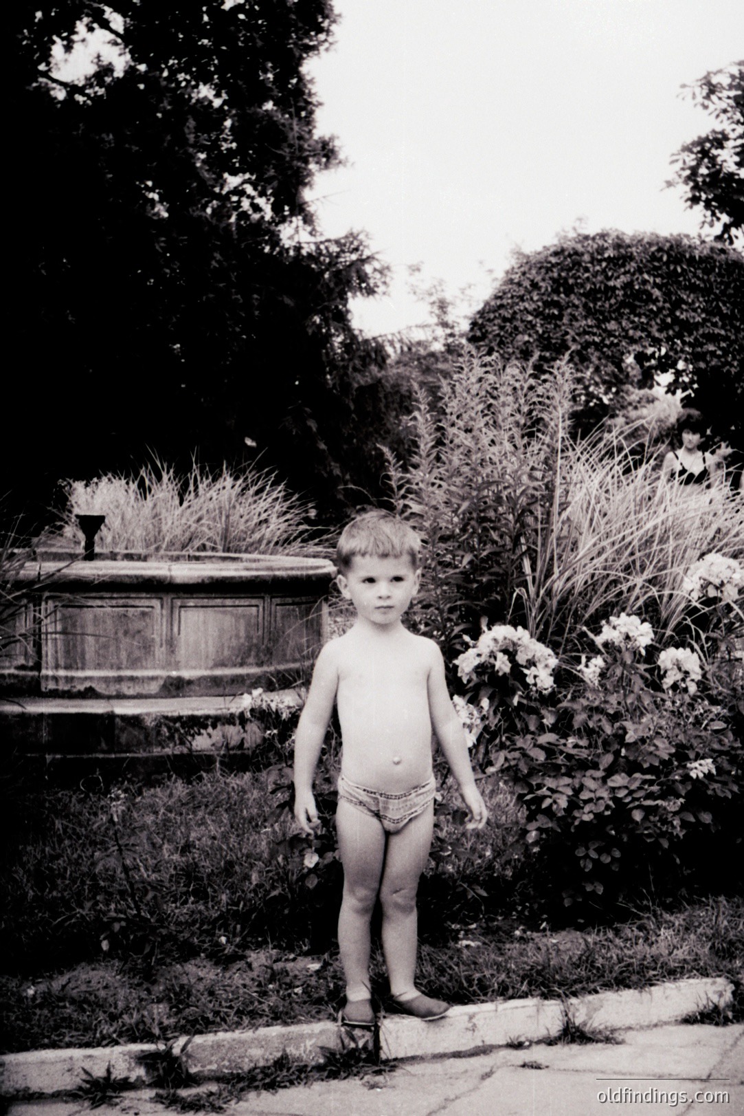 A young boy stands barefoot, partially obscured by foliage, in a formal garden setting. He wears only briefs. A woman's back is visible in the blurred background. Likely a candid snapshot from the mid-20th century. High archival and design value for portraying childhood.