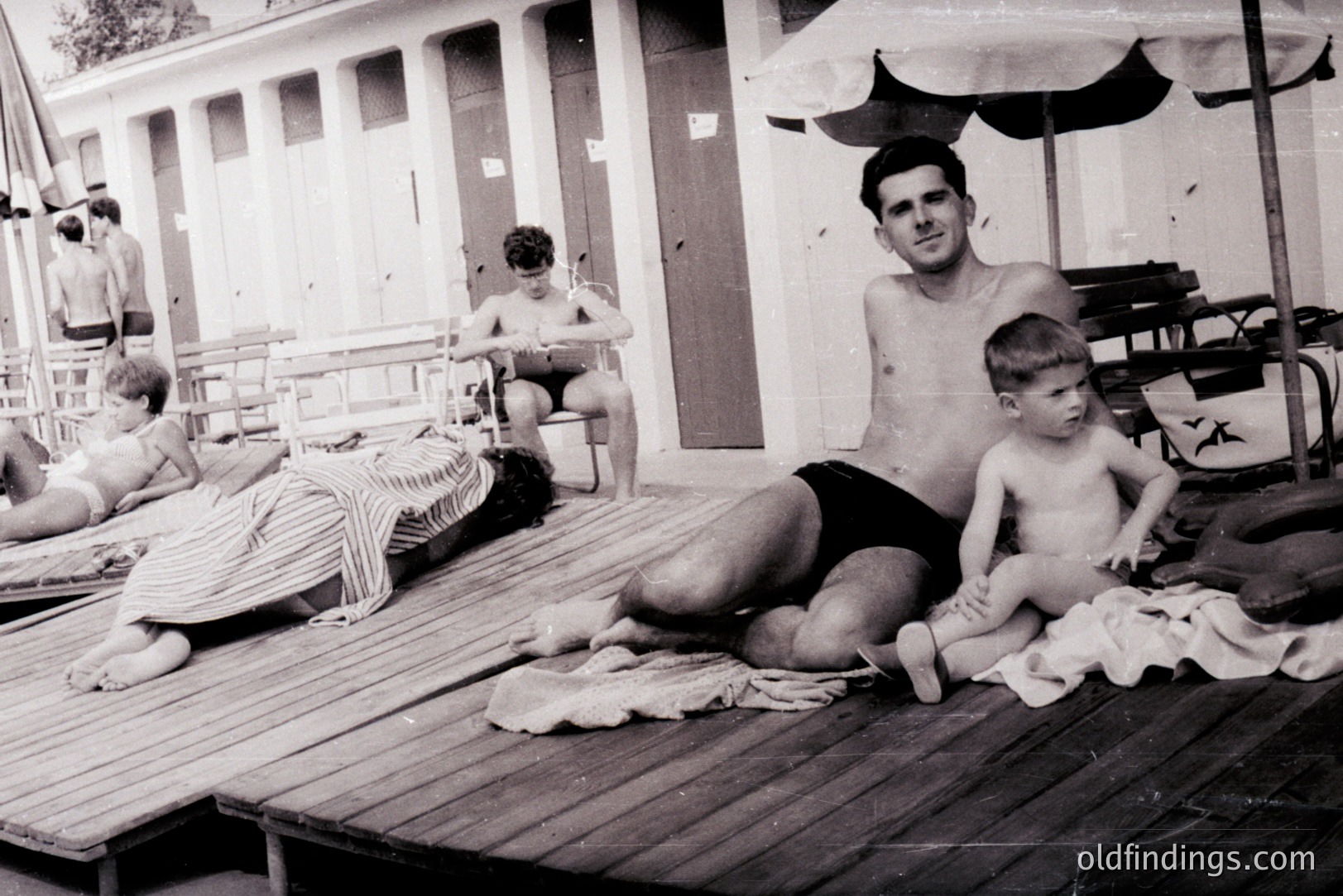A candid poolside scene: A man and young boy relax on a wooden deck, while a young woman sunbathes nearby. Architectural details suggest a mid-century resort or public pool setting. A classic striped beach umbrella provides shade. Likely 1950s-1970s.