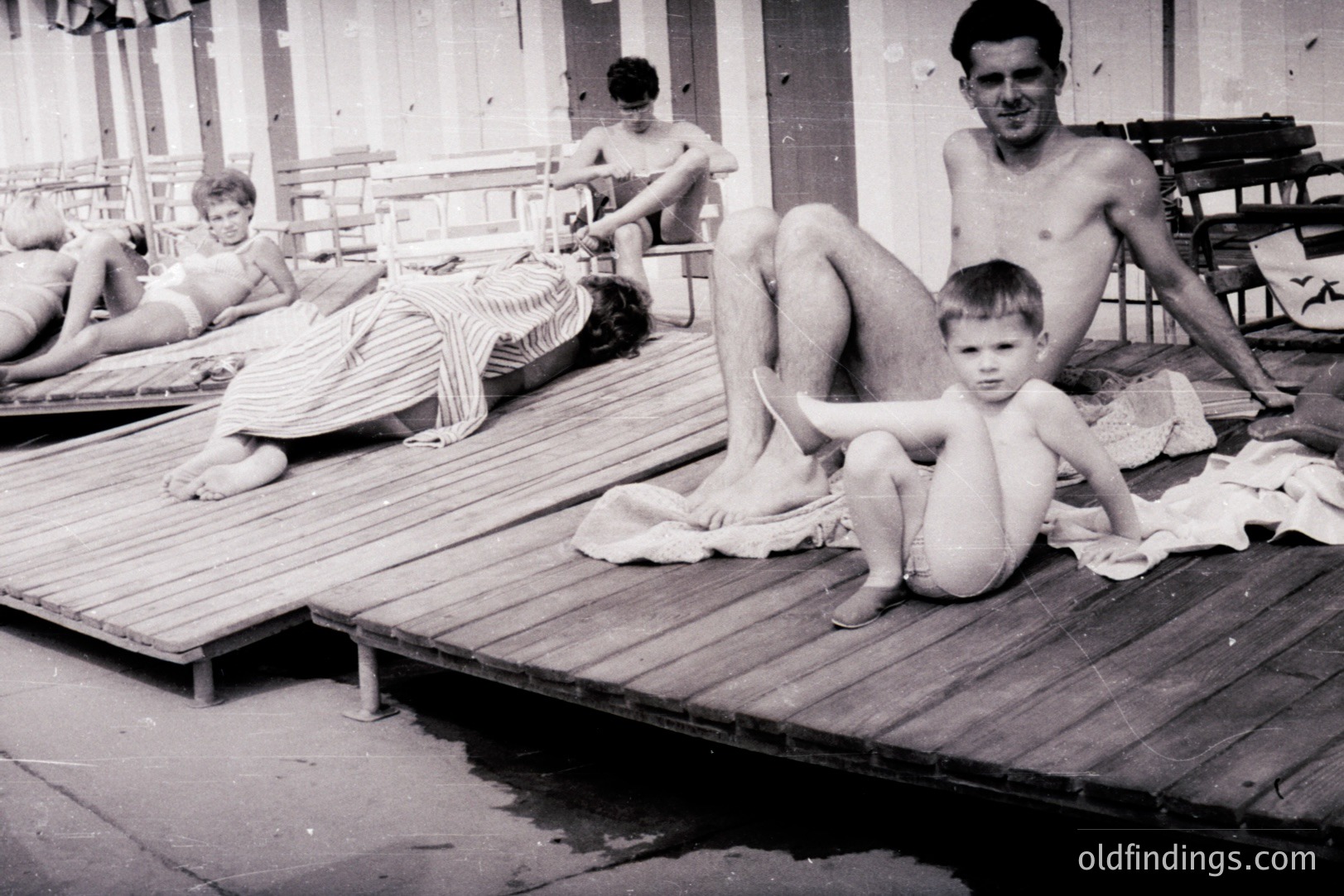A vintage monochrome photo captures a family scene on a wooden boardwalk. A man sits with a young boy, both nude; a woman lies on a nearby lounge chair. The background reveals striped awnings & simple wooden furniture suggesting a seaside resort. Likely 1960s or 1970s.
