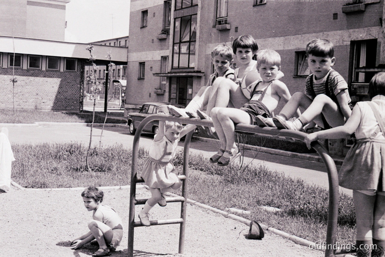Group of children play on a seesaw in a concrete courtyard, likely a Soviet-era housing complex. The architecture suggests Eastern Europe, possibly 1960s-1970s. Boys wear shorts & some have suspenders; a girl wears a dress. Playground equipment shows signs of use. A vintage car is visible.