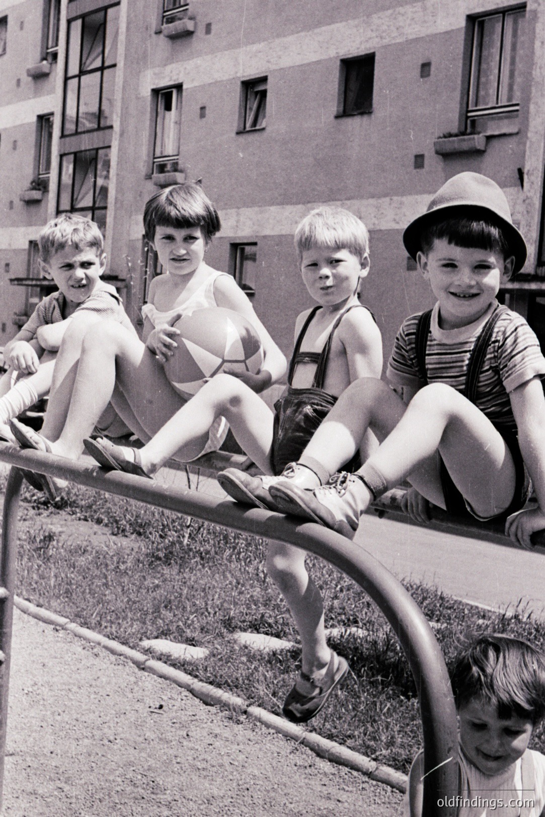 Four boys playfully perch on a metal playground structure in front of a block of apartments. Distinctive clothing – overalls, hats, sandals – suggests a mid-20th century, likely 1960s, Eastern European setting. A basketball is held by one boy. Image valuable for documenting childhood and urban design.