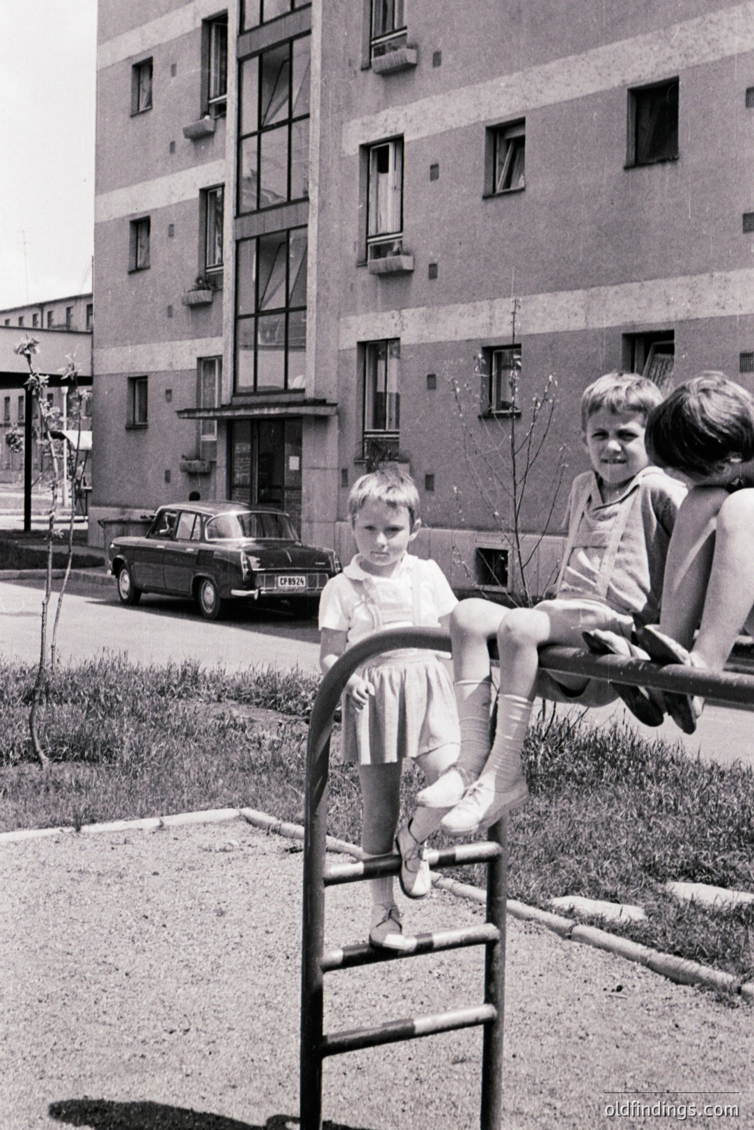Two children sit and pose on a metal playground structure, a typical example of mid-century recreational design. A Soviet-era sedan is parked on the street in front of a large, blocky apartment building. Likely Eastern Europe, 1960s-1970s. A study in socialist architecture & childhood.