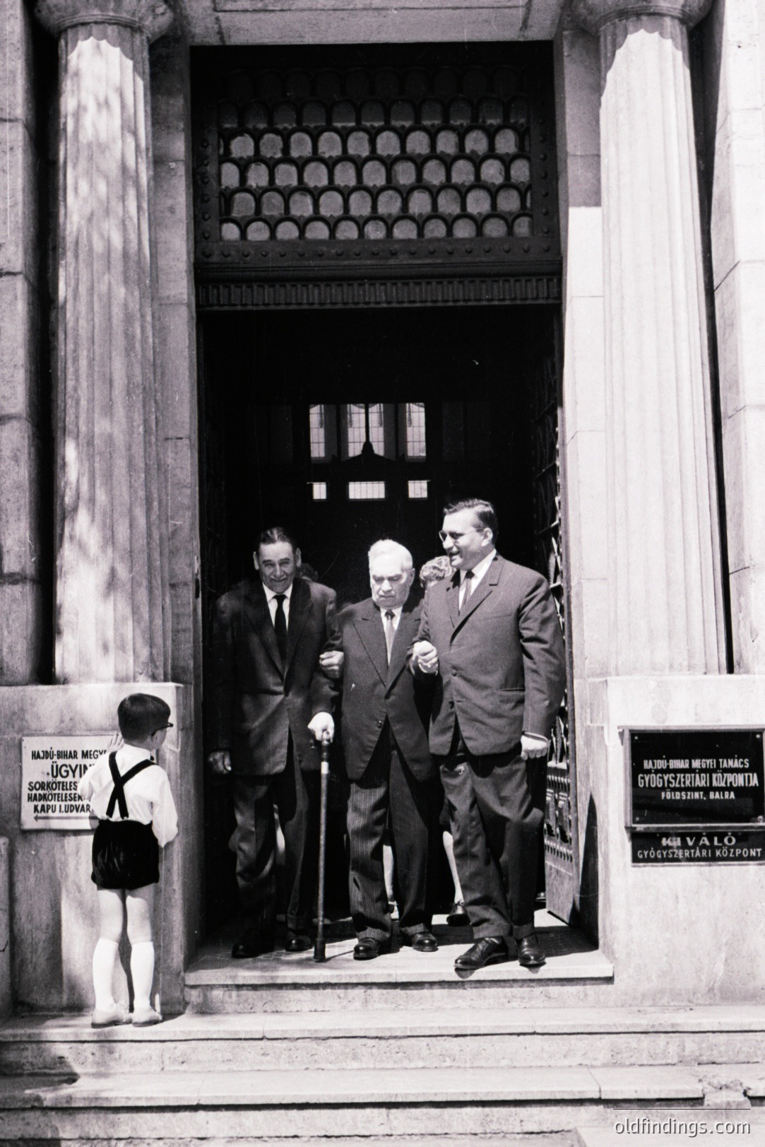 Formal black and white photo of three men in suits entering an ornate building. A young boy in short pants and suspenders stands on the steps. Signage visible in Hungarian. Architectural details suggest a European, possibly Eastern European, locale. Likely 1960s-1970s era.