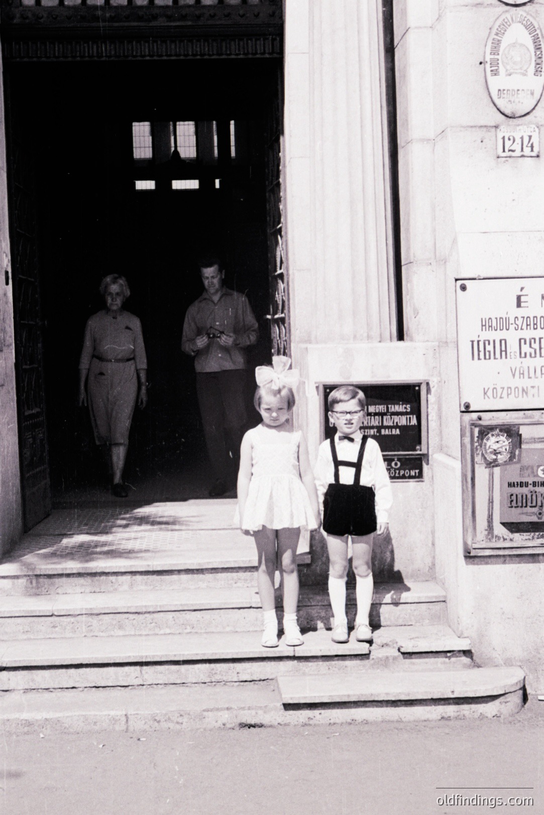 Two young girls, one in a dress & bow, the other in pinafore & suspenders, stand on stone steps. Adults exit a building behind. Signage suggests a location in Hungary. Likely mid-20th century, possibly 1950s-1960s, showcasing period fashion. Historic childhood portrait.