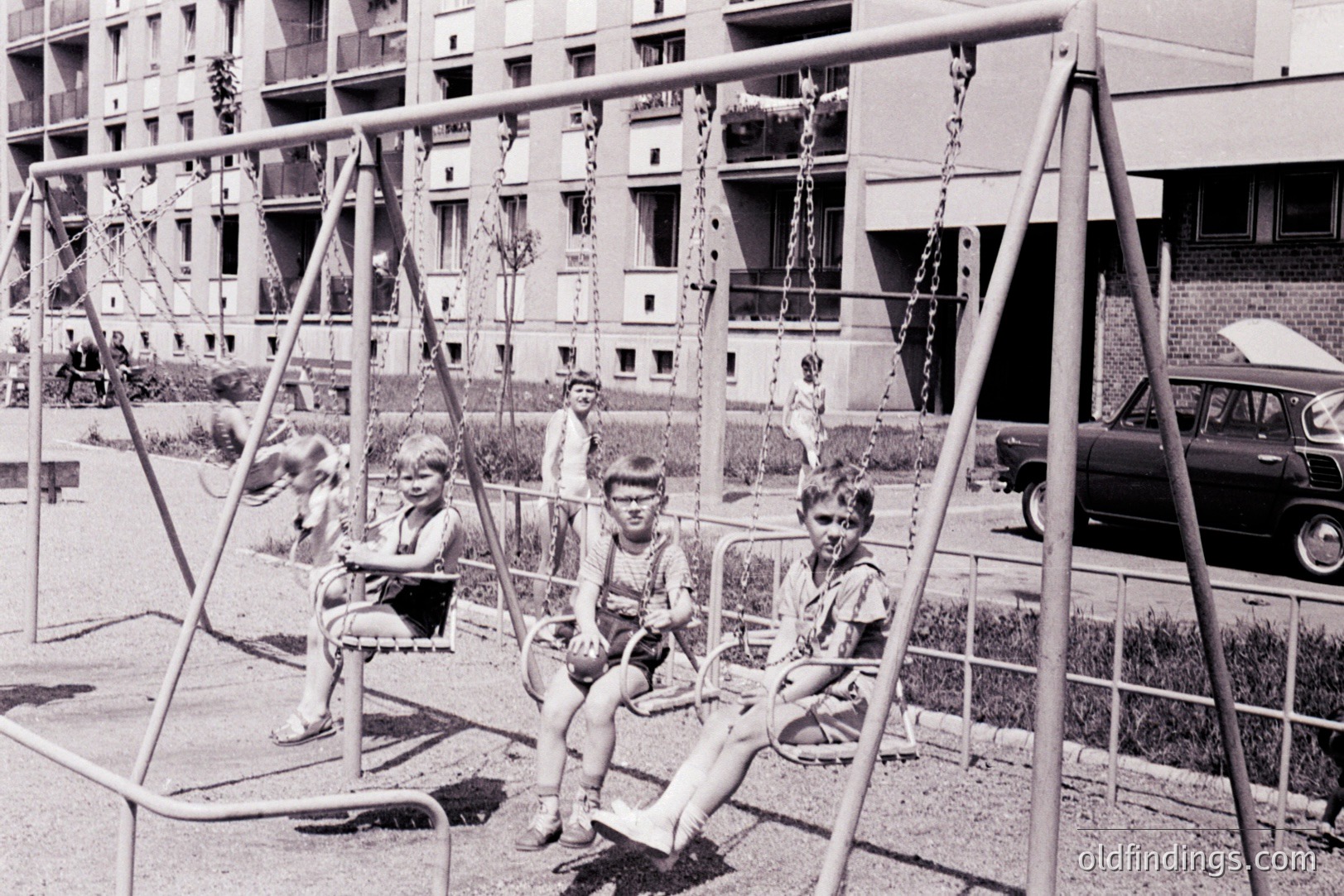 Three boys seated on swings in a public playground. Housing block architecture visible in the background, indicative of post-war social housing. A vintage car is parked nearby. Likely 1960s, Eastern Europe. Possible candid snapshot of childhood leisure.