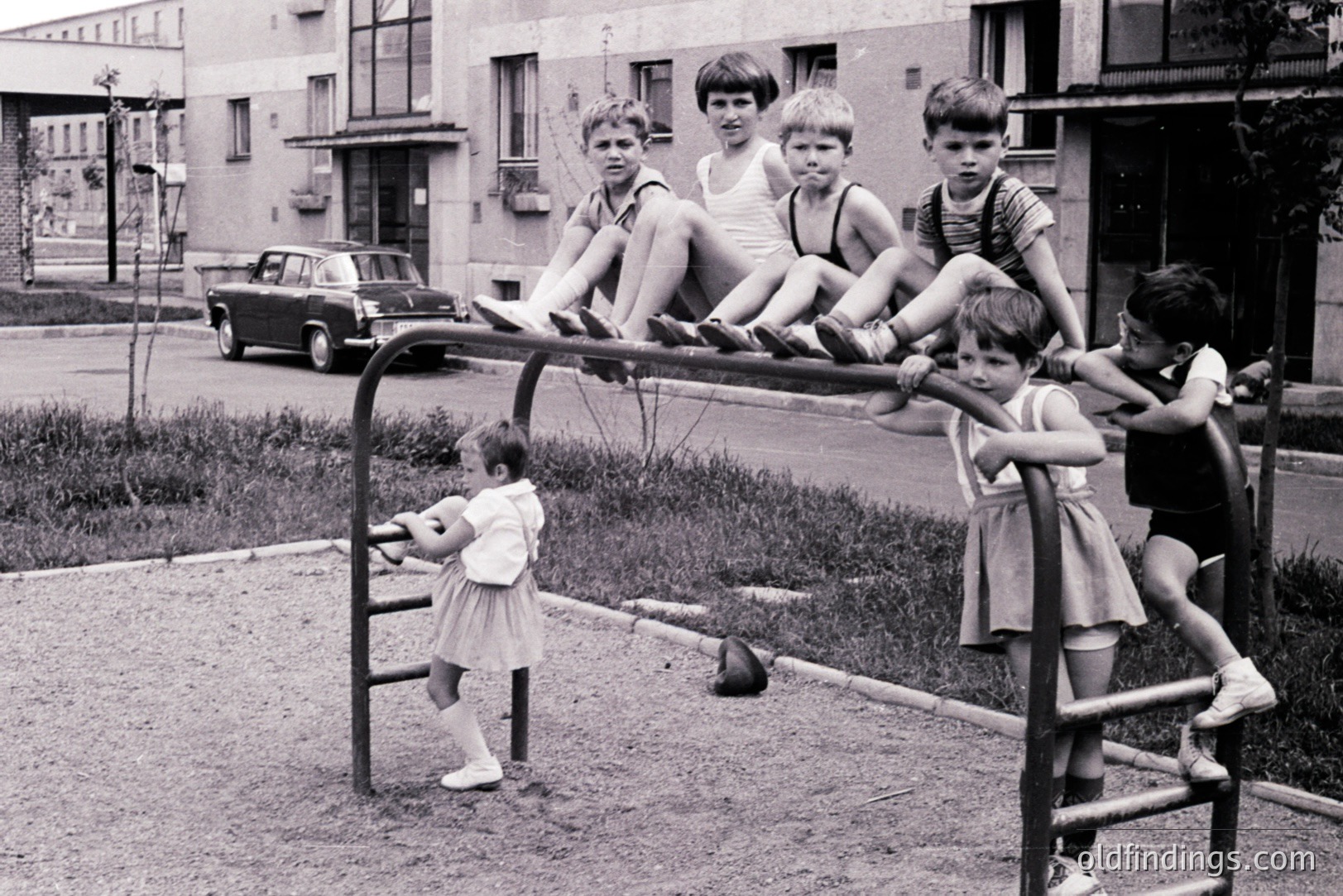 Group of boys perched on a gymnasium structure, a girl below. Mid-century apartment complex backdrop; a dark sedan parked nearby. Likely a Soviet-era playground, potentially Eastern Europe. Capture of childhood recreation, simple leisure. Architectural context highlights post-war housing.