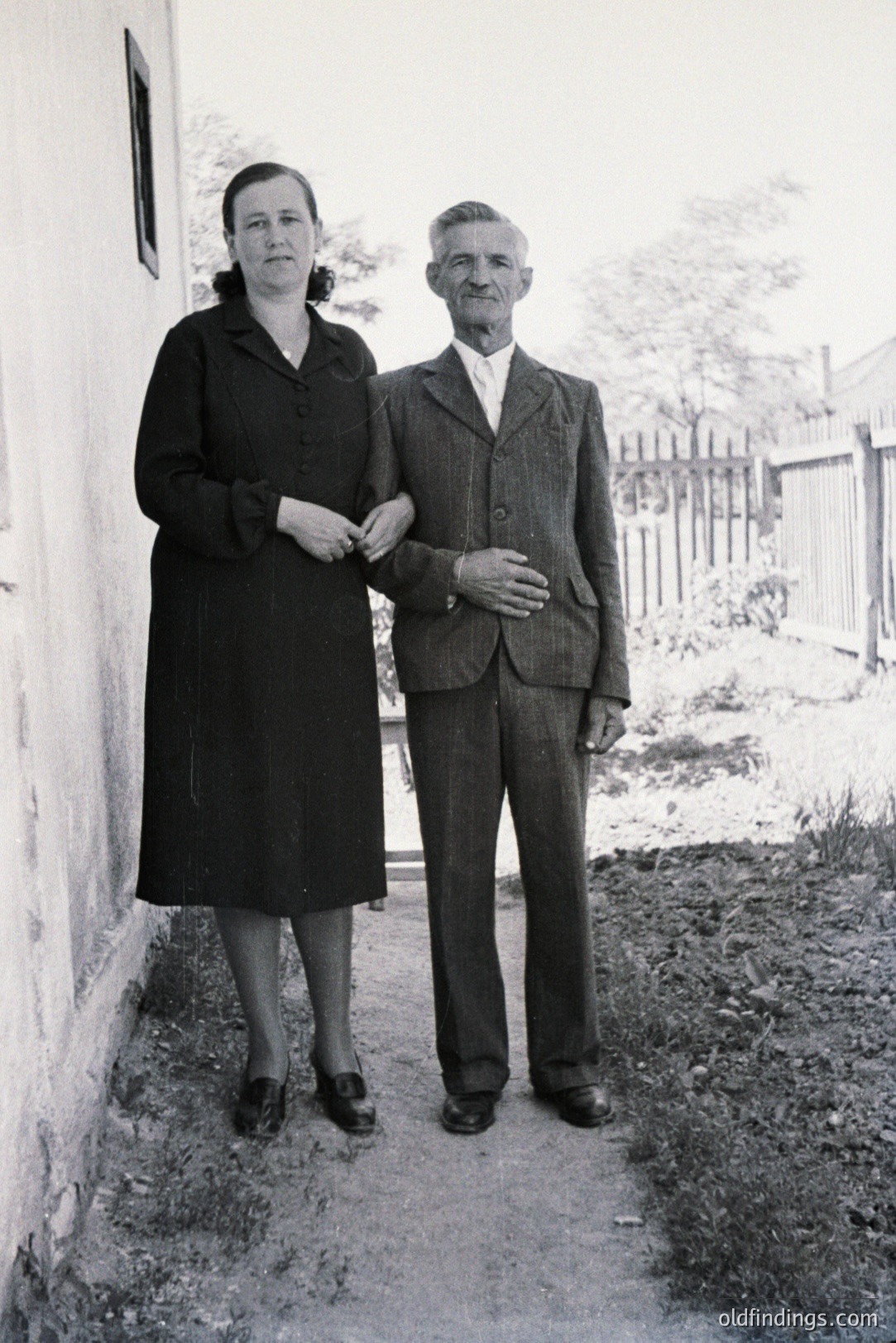A formally dressed elderly couple stands outdoors, leaning against a weathered wall. The woman wears a dark, knee-length dress and shoes; the man, a three-piece suit and tie. The setting appears to be a rural area with a wooden fence and simple architecture. Likely taken in the 1950s or 1960s. Valuable for family history or period design reference.