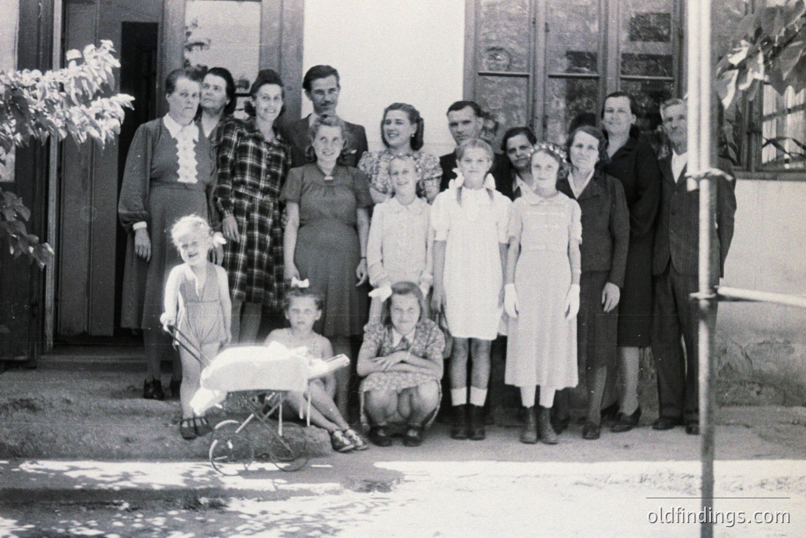 A formal group portrait; ten individuals and three children pose outside a building with a covered entryway. Clothing suggests a 1930s-1940s timeframe. The children are posed near a wheelbarrow. Likely a family or community gathering. Black and white print.