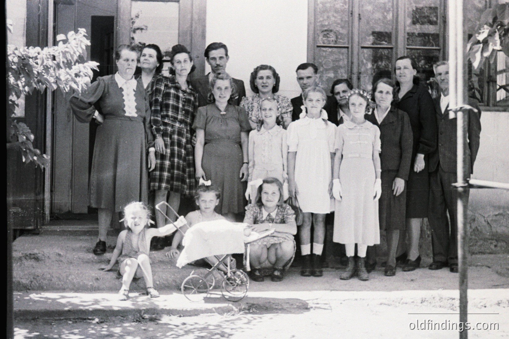 A large family group poses outdoors, likely in front of a home or institutional building. Women in mid-century dresses and men in suits occupy the frame. A baby lies in a pram. Architectural details suggest a European location. Likely 1940s-1950s. Potential value for family history research.