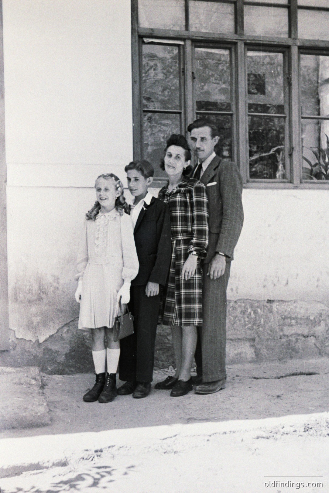 A family portrait: a girl, boy, and parents stand before a building with visible architectural details. The girl wears a dress & boots, boy in a suit with a briefcase. Parents in plaid & tweed. Likely 1940s-50s, possibly rural setting. Strong composition, potential for historical/design reference.