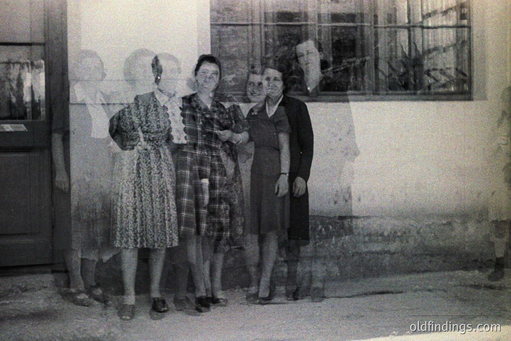 Four women stand formally outdoors, posed against a building’s exterior. Dresses with patterns and modest necklines suggest a 1940s or 50s era. Visible architectural details include a glass-paned door and textured plaster. The photo's layered effect is unusual, potentially from double exposure.