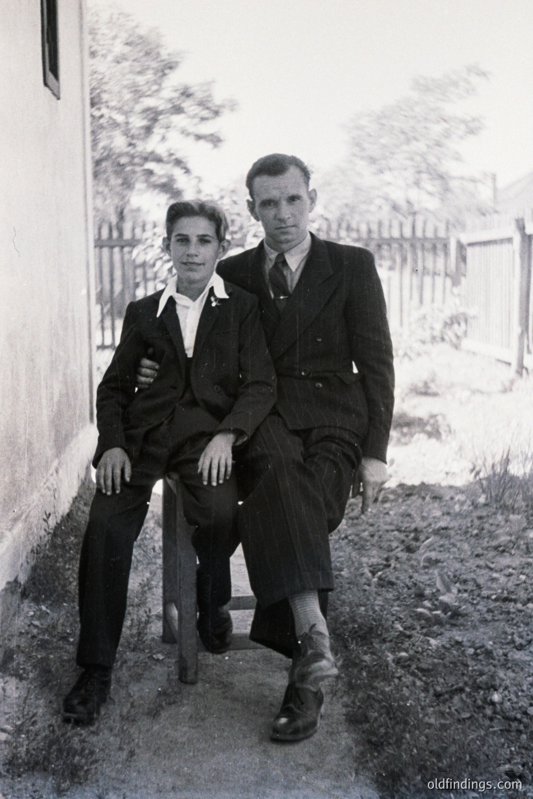 Formal portrait of a man and a young man seated outside a building, likely a home. The men wear dark suits, ties, and polished shoes; a classic, slightly austere style. The backdrop shows a simple fence and vegetation. The photograph’s grainy texture suggests the 1940s or 1950s.