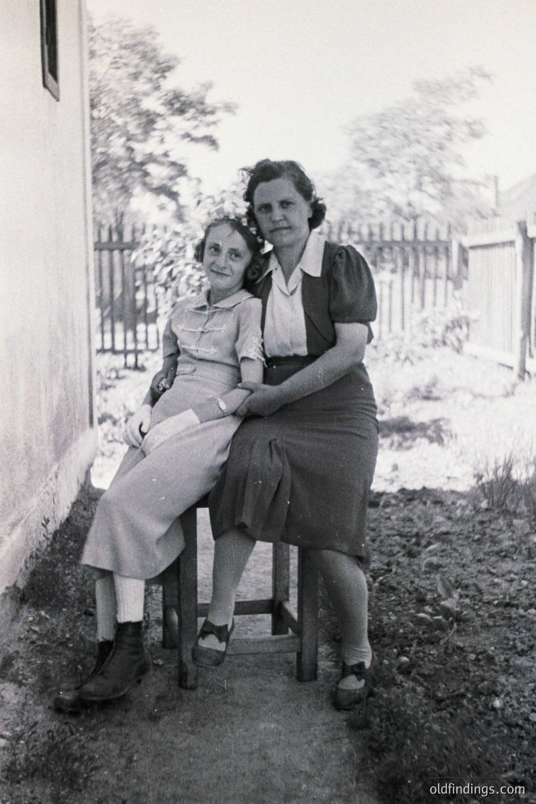 Two women sit outdoors, posed near a plain, stucco wall. Both wear modest, 1940s-style dresses and sturdy shoes. One woman’s hair is dark and curly. A simple wooden fence and sparse vegetation are visible in the background. Likely a family portrait.
