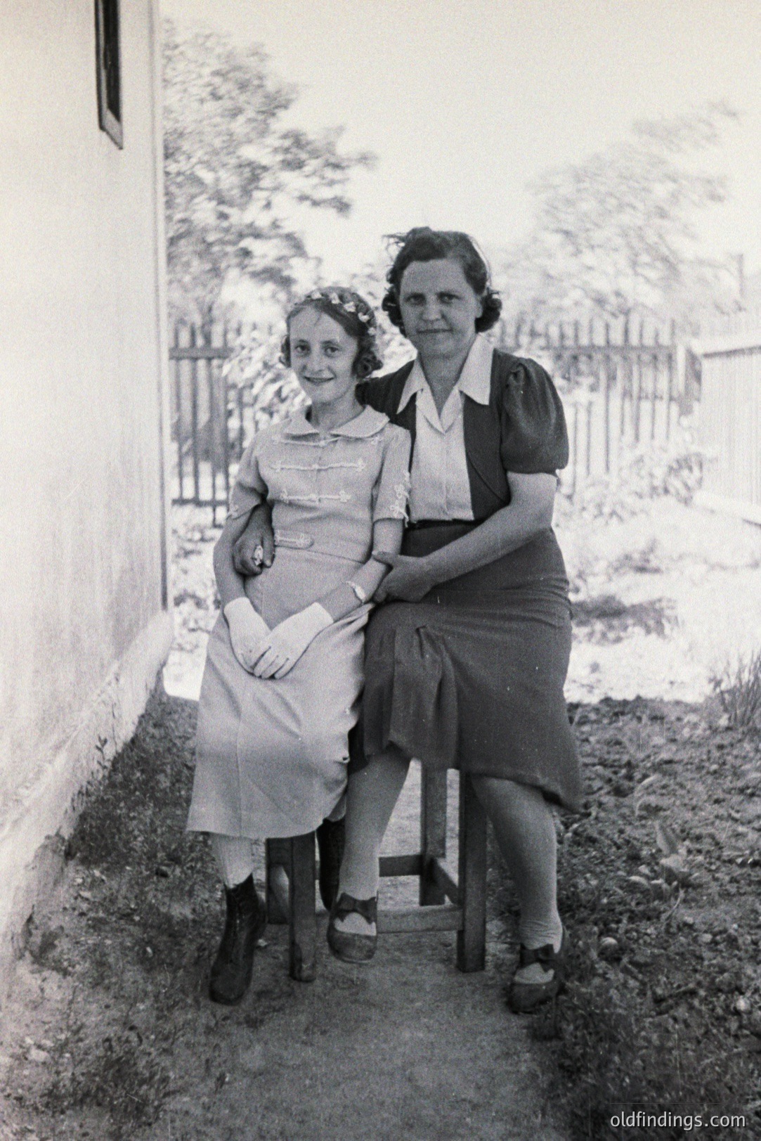 A young woman in a dress and older woman in a collared shirt sit side-by-side on simple stools. The setting appears to be a courtyard or garden with a weathered wall and fence. Likely a family portrait, possibly post-war era (1940s-50s). Simple attire suggests modest circumstances. The lighting is natural, highlighting textures.