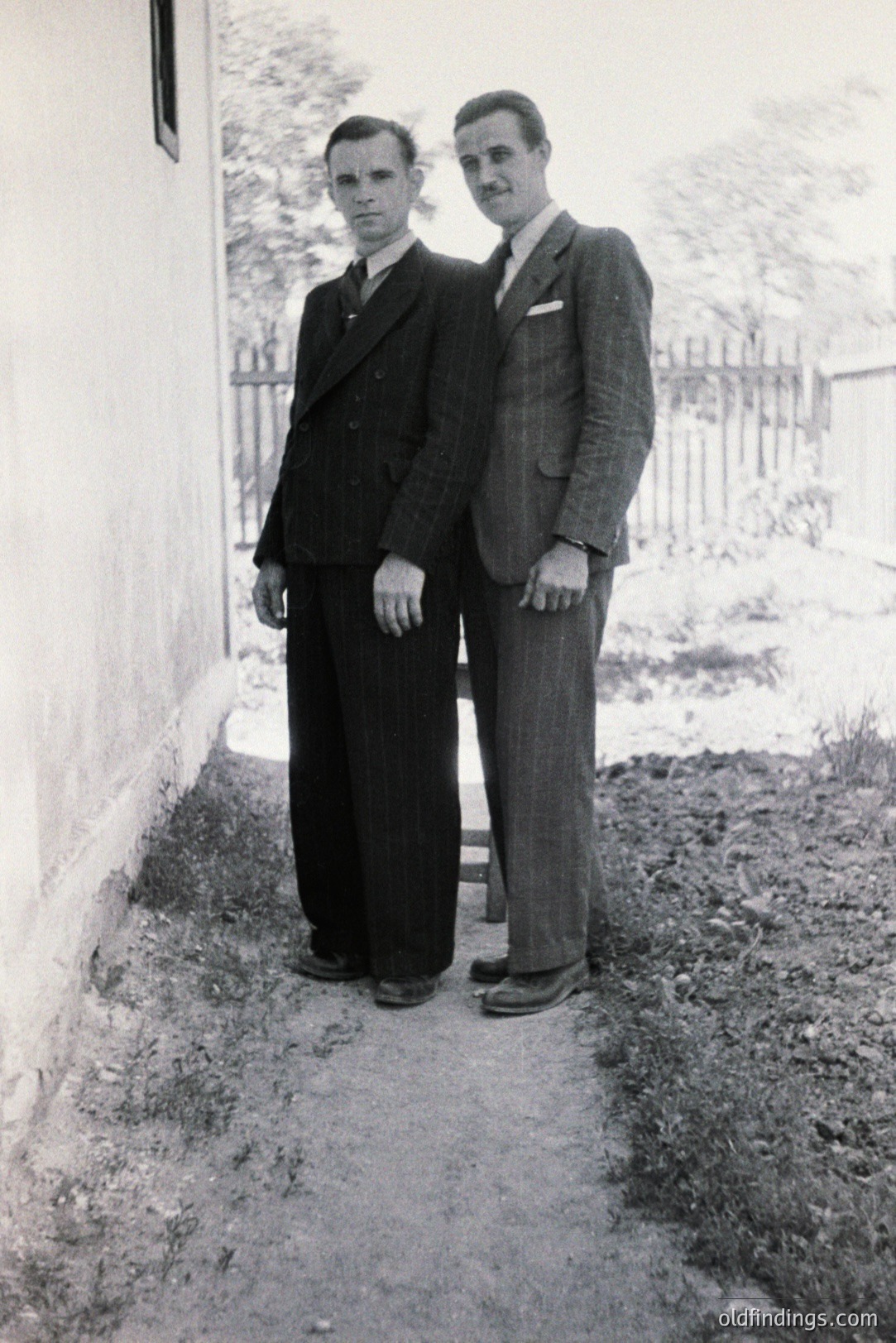 Two men stand beside a stone wall, formally attired in 1940s-style suits. The man on the left wears a dark double-breasted suit; the other, a plaid suit. A dirt path and rustic fence are visible. Likely a portrait taken outdoors. Photographic value: family history, vintage fashion reference.