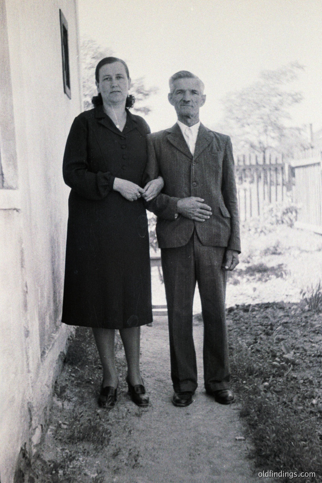 A formally dressed older couple stands near a simple whitewashed building. The woman wears a dark, knee-length dress and Mary Jane shoes. The man is in a three-piece suit and tie. Likely a portrait from the 1950s-1970s, possibly Eastern Europe. Reflects post-war austerity and traditional portraiture.