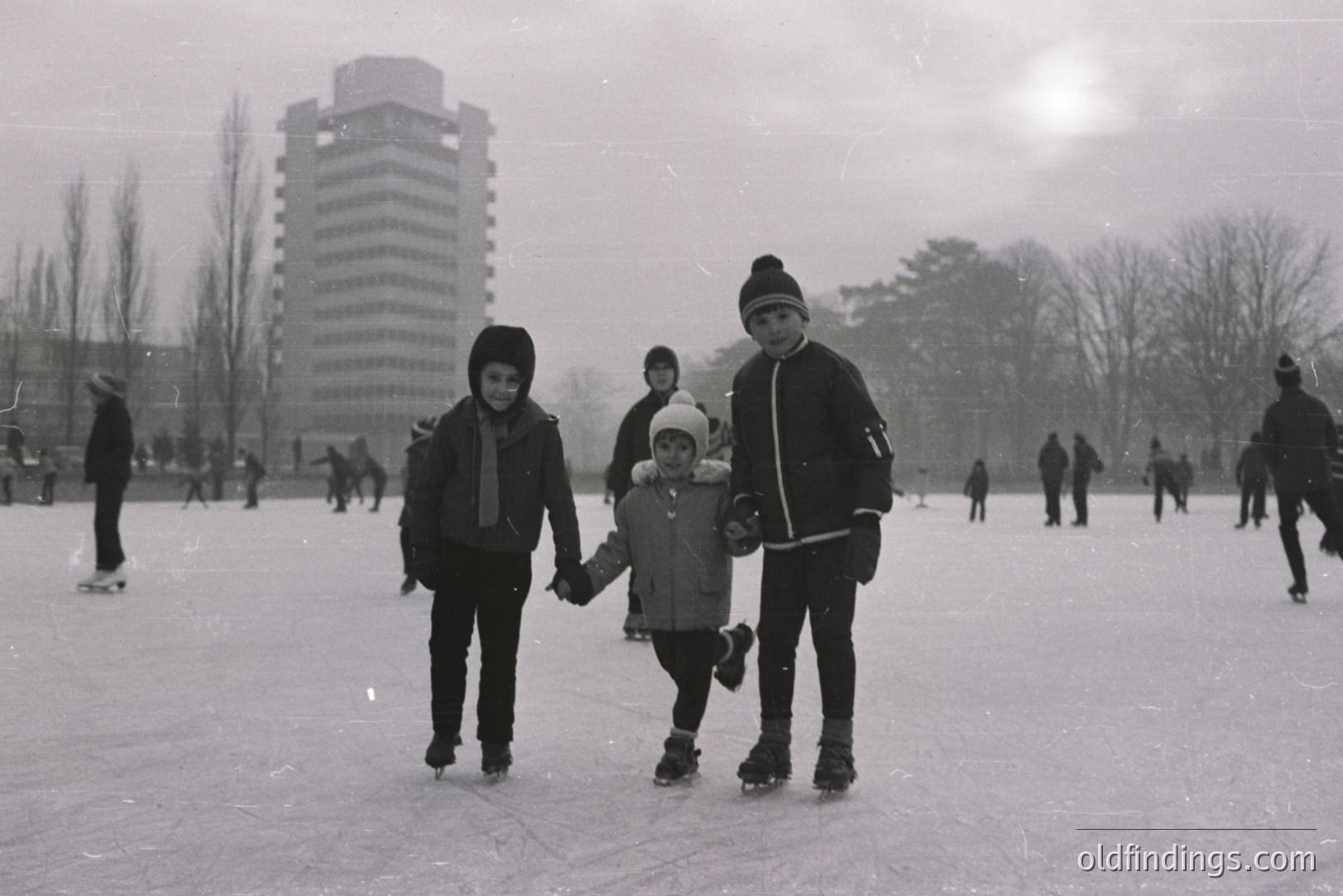 Three children ice skating on a frozen pond or lake, linked hands. A tall, brutalist apartment building rises in the background. Appears to be a public space with numerous other skaters. Likely 1960s-1970s, Eastern Europe or similar climate. Winter scene, overcast sky.