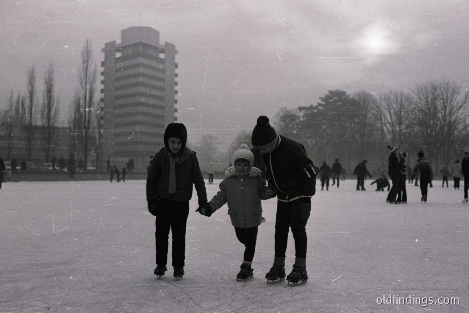 Monochrome image showing a man, presumably a father, supporting a young child learning to ice skate. A second child stands nearby. A tall, modernist apartment building is visible in the blurred background. Likely 1960s-1970s, captured during a period of natural ice formation. skating