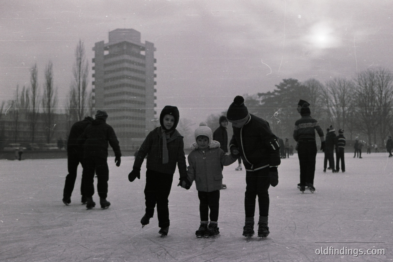 A group of children ice skating on a frozen pond, seemingly outdoors. A taller figure appears to be assisting the younger skaters. A tall, modernist apartment building is visible in the background. Likely a community recreation scene, circa 1960s-1970s. Winter leisure activity.