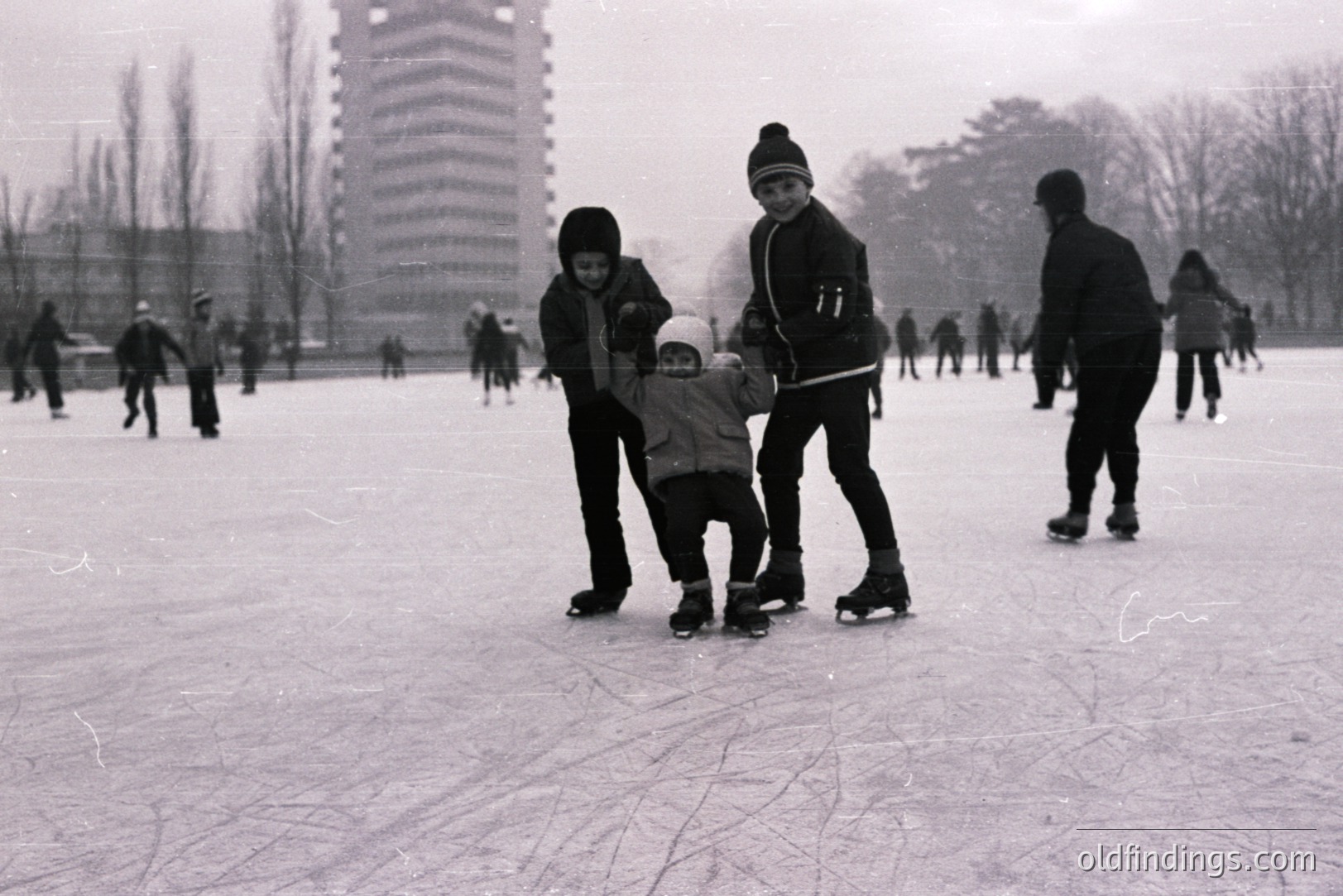A young boy, assisted by two older children, learns to ice skate on a frozen pond. Background shows other skaters and a tall, modernist apartment building. Appears to be a recreational winter scene, likely 1960s or 70s. The grainy quality is characteristic of older film stock.