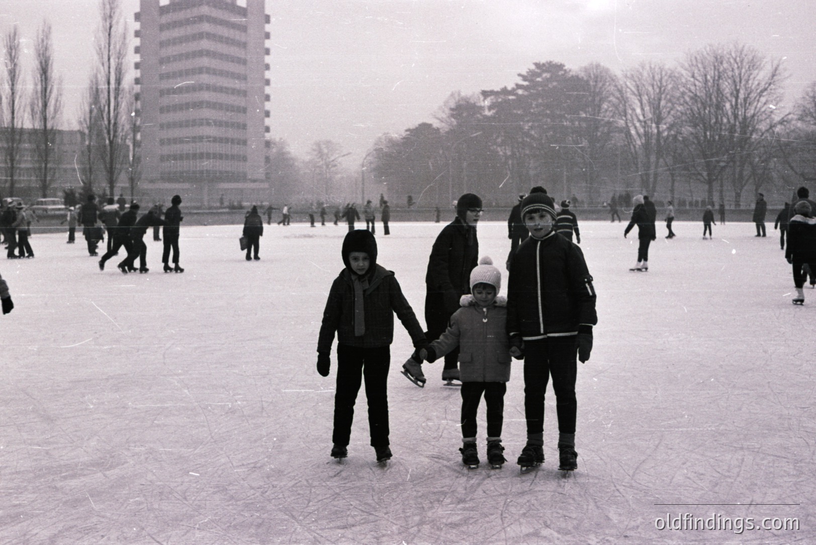 Two children, holding hands, stand on a frozen pond/lake amidst a crowd of ice skaters. A tall, modernist apartment building rises in the background. The monochrome image evokes a wintry scene. Likely 1960s-1970s. A candid moment of childhood recreation.