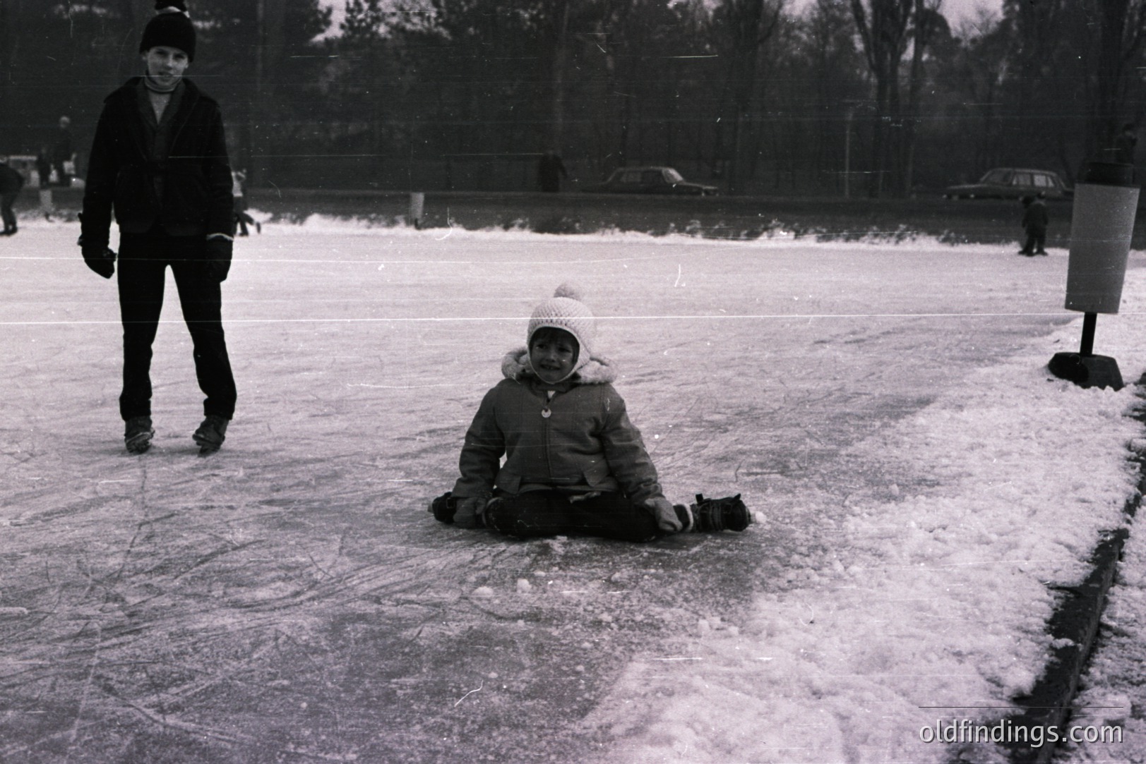 Black & white photo showing a child seated on a snowy outdoor ice rink. A second individual is visible in the background, skating. Likely a public recreation area, 1960s-1970s aesthetic. Snow-covered ground, winter scene. Candid moment, documenting leisure activity. Potentially valuable for historical recreation or design.