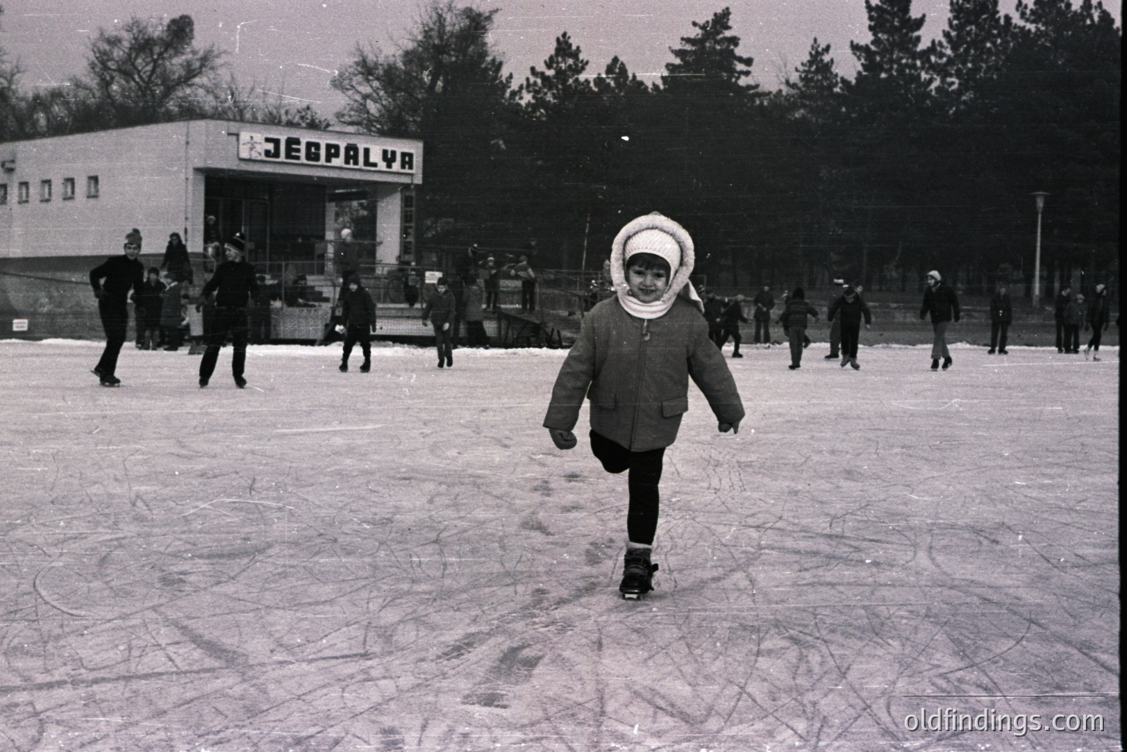 A young girl, bundled in a winter coat and hat, stands poised on skates on a frozen pond. Several other skaters populate the scene, some visible near a building with Cyrillic signage ("Zebalya"). Likely a recreational area in Eastern Europe, possibly Bulgaria. Appears to be mid-century, ~1960s. A charming glimpse of leisure time.