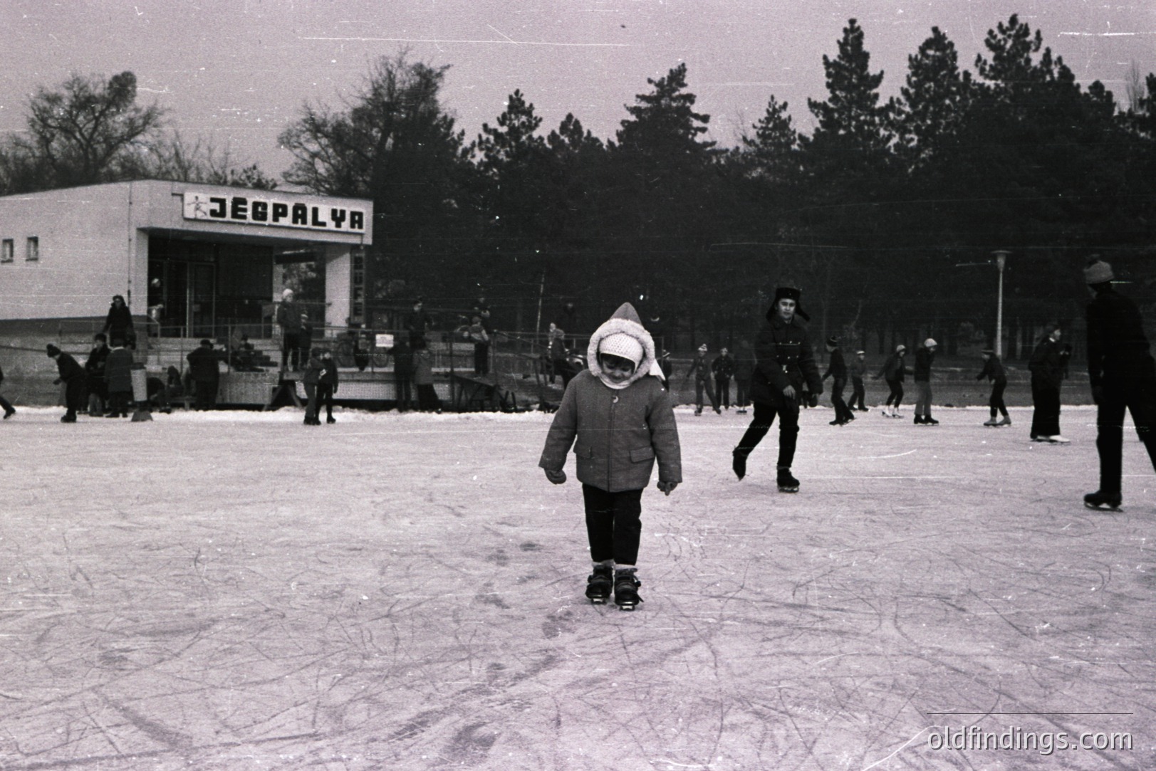 A monochrome image of an outdoor ice skating rink, likely in Eastern Europe. A child in a hooded coat stands center frame, seemingly preparing to skate. Several other skaters populate the scene. Building signage reads "LEGPALYVA". Architectural style suggests mid-century Soviet influence. Appears to be a public recreational space.