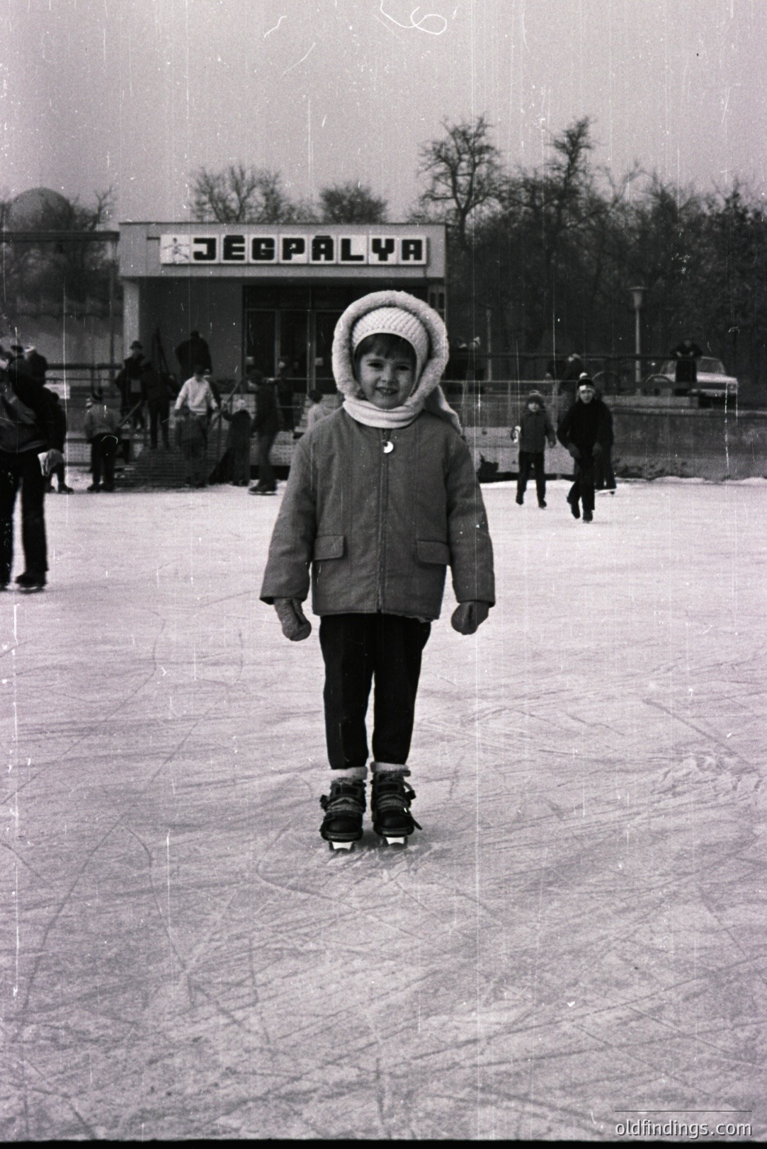 A young child, bundled in a warm coat & hat, stands confidently on an outdoor ice rink. "ZEBPALYA" signage visible in the background, suggesting a recreational facility. Likely 1960s-70s, Eastern Europe. Several other skaters present. A candid moment.