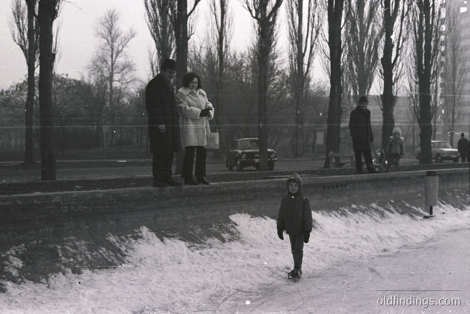 A monochrome street scene depicts a young boy in a dark coat walking along a snow-covered sidewalk. In the background, a couple in winter coats and a man with a child in a stroller observe. Vintage car models suggest a 1960s or 70s setting. Likely Eastern European, possibly urban.