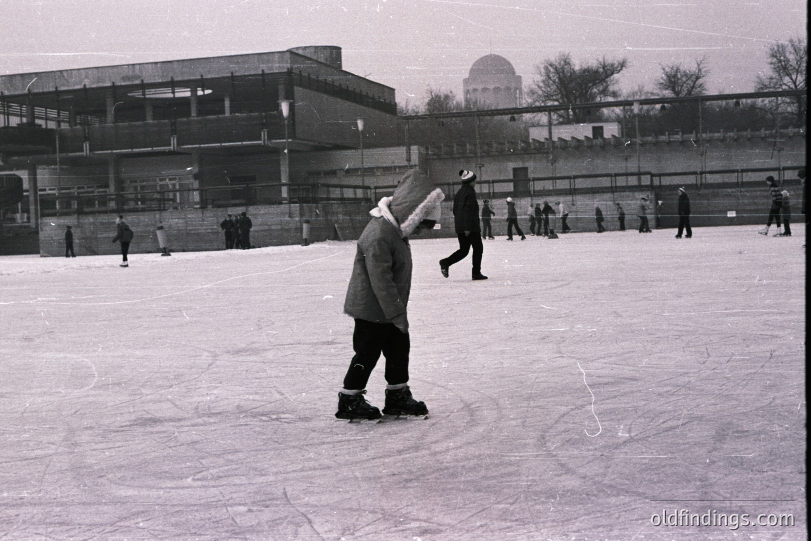 B&W image captures a public ice rink scene. Several figures skate and walk across the ice. A large, modern building with a curved roof looms in the background. Likely a recreational facility, possibly 1960s or 70s. Atmospheric, candid moment of winter leisure.
