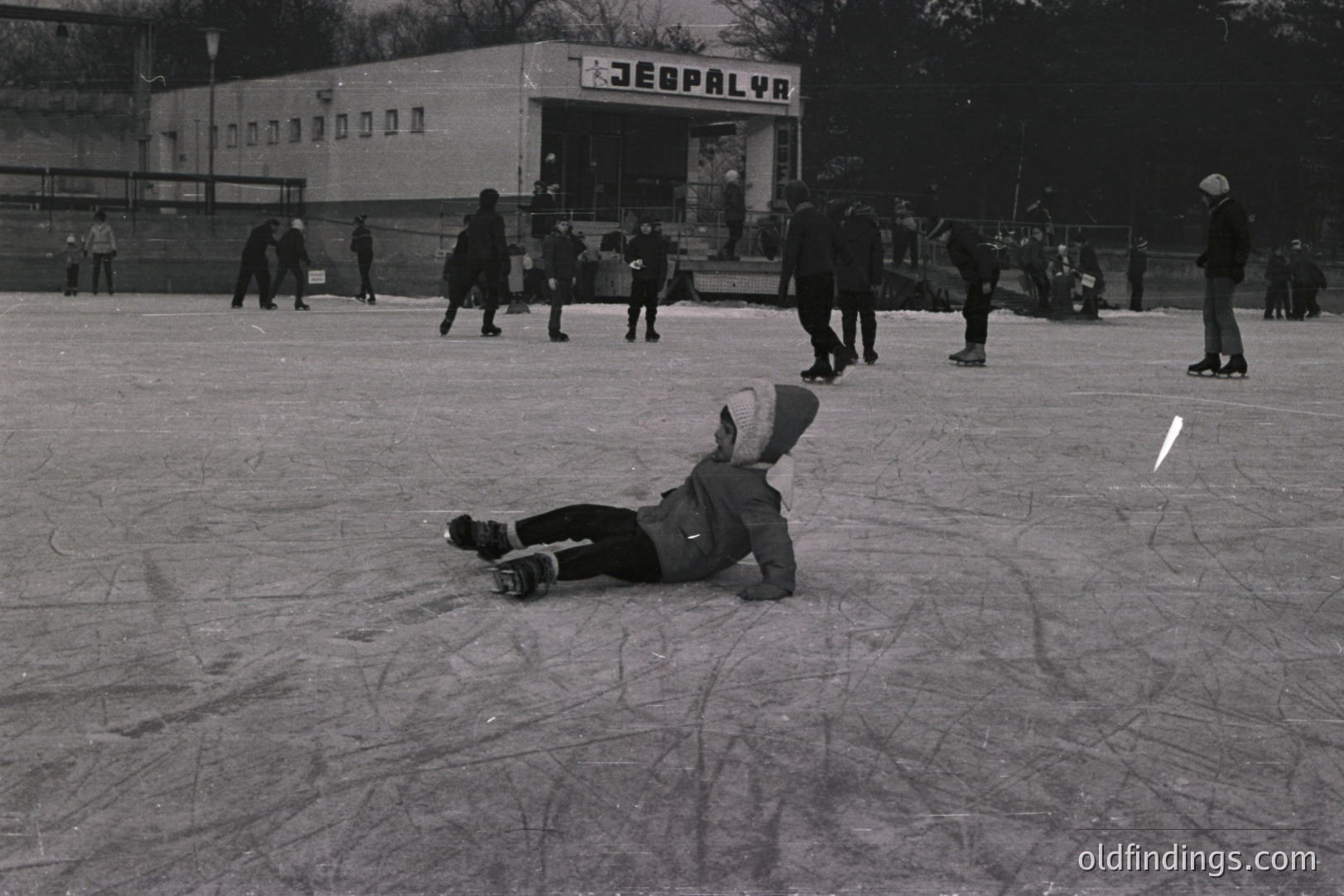 A child in winter clothing lies prone on a frozen outdoor ice rink, surrounded by other skaters. The "JEBPLYR" sign of a building is visible in the background. A typical recreation scene, likely from the 1960s-1970s.