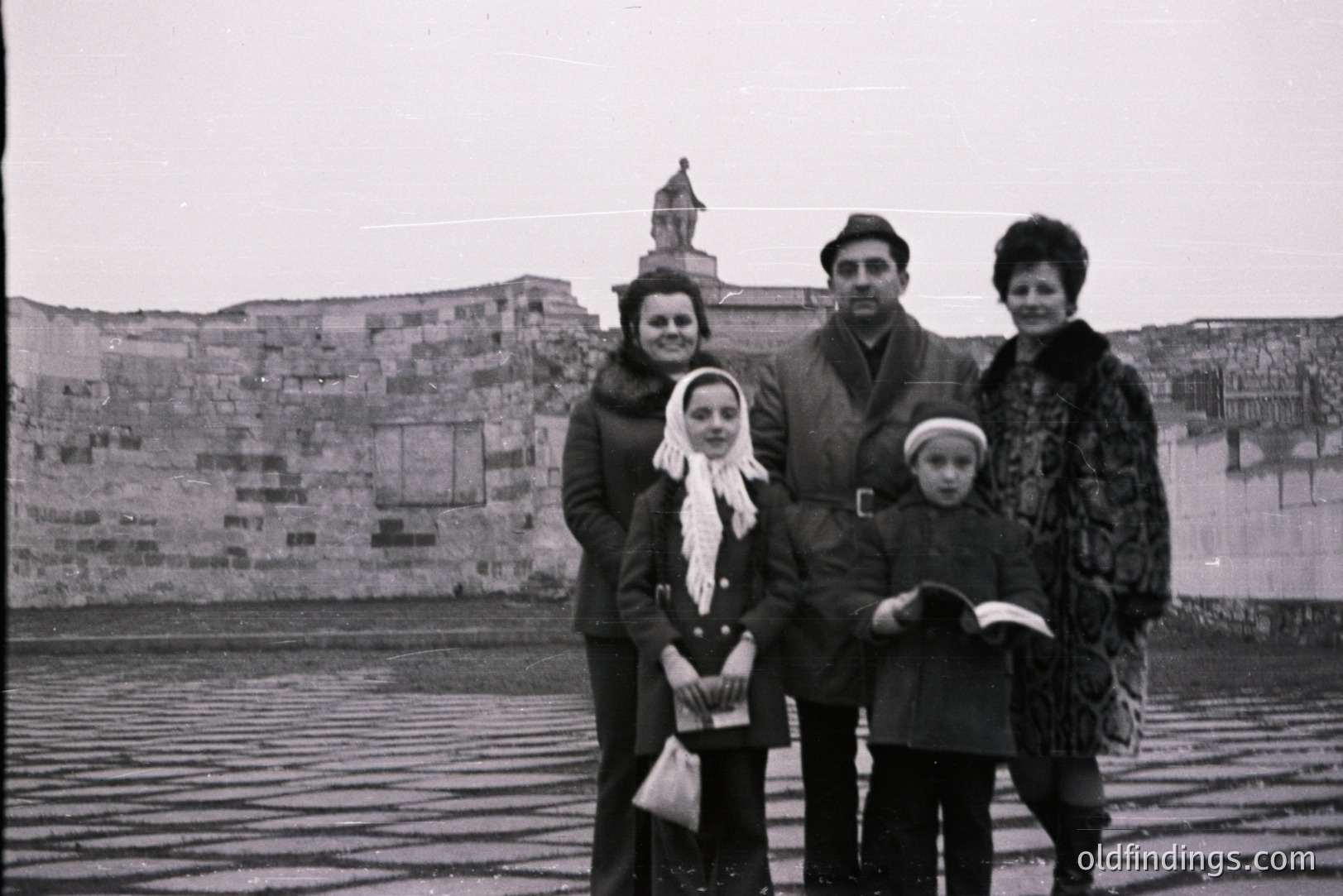 A family portrait from the 1960s or 70s. Four people stand outdoors before a stone wall topped with an ornate architectural structure and a statue. The family, likely on vacation, is dressed in era-appropriate clothing. A youthful, candid feel is present.
