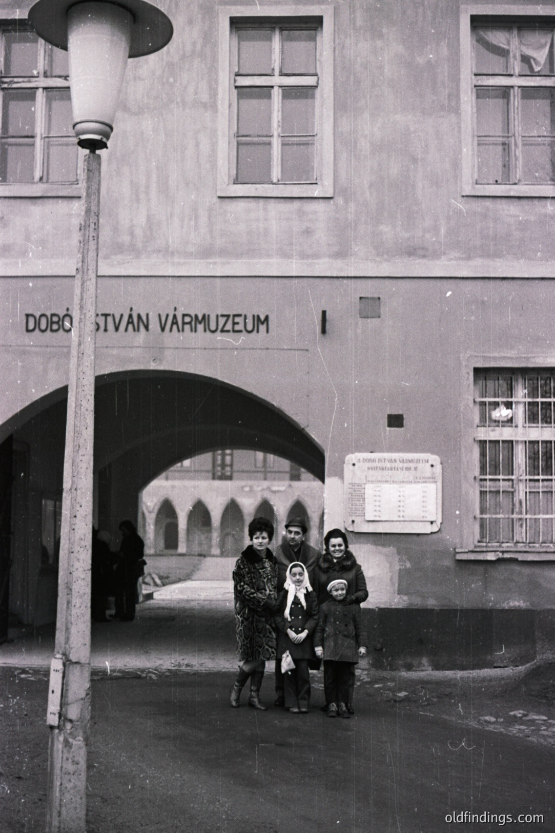 A family stands in a stone archway before a building with signage reading "Dobó István Vármúzeum." Likely 1960s or 70s, showing mid-century fashion, including a fur collar. The museum suggests a historical or cultural site. A simple, functional architectural style.
