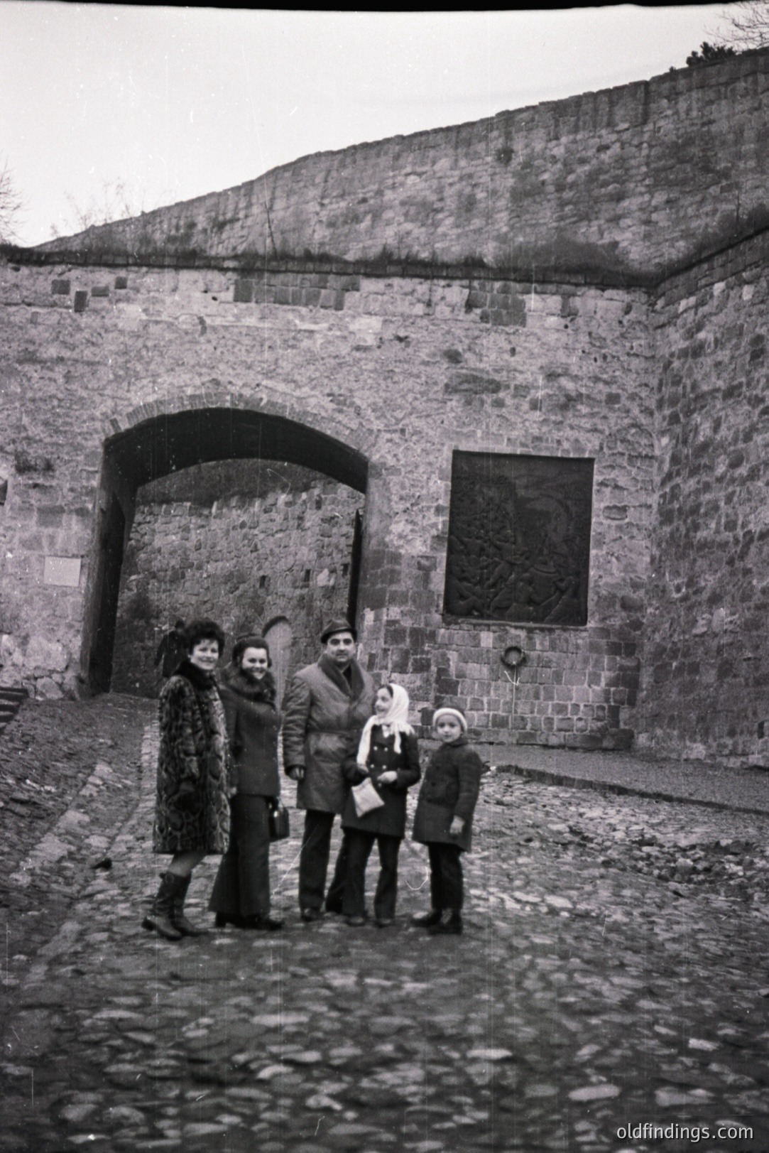 Family portrait before a stone archway, likely a fortress gate. Individuals are dressed in 1970s outerwear & bell-bottoms. A large stone relief depicting a coat of arms is centered above the arch. Paved courtyard and stone wall create a fortress-like aesthetic. Possible travel/tourism documentation.