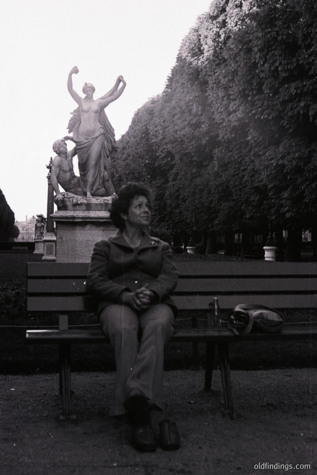 A seated individual wearing a dark jacket and corduroy trousers occupies a park bench before a monumental statue. Dense foliage fills the background. Likely 1970s style. Appears to be a candid street photograph, potential for documentary or design use. Shoes are visible on the bench beside a small bag.