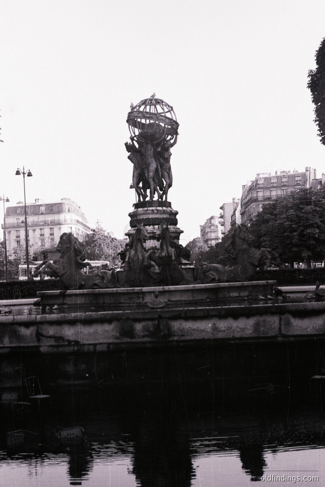 A monumental fountain featuring a sculpted figure holding a globe, set within an elaborate basin, is seen reflected in water. Background shows a cityscape with architectural details and trees. Likely a public space or park. Photographic style suggests a mid-20th century aesthetic.