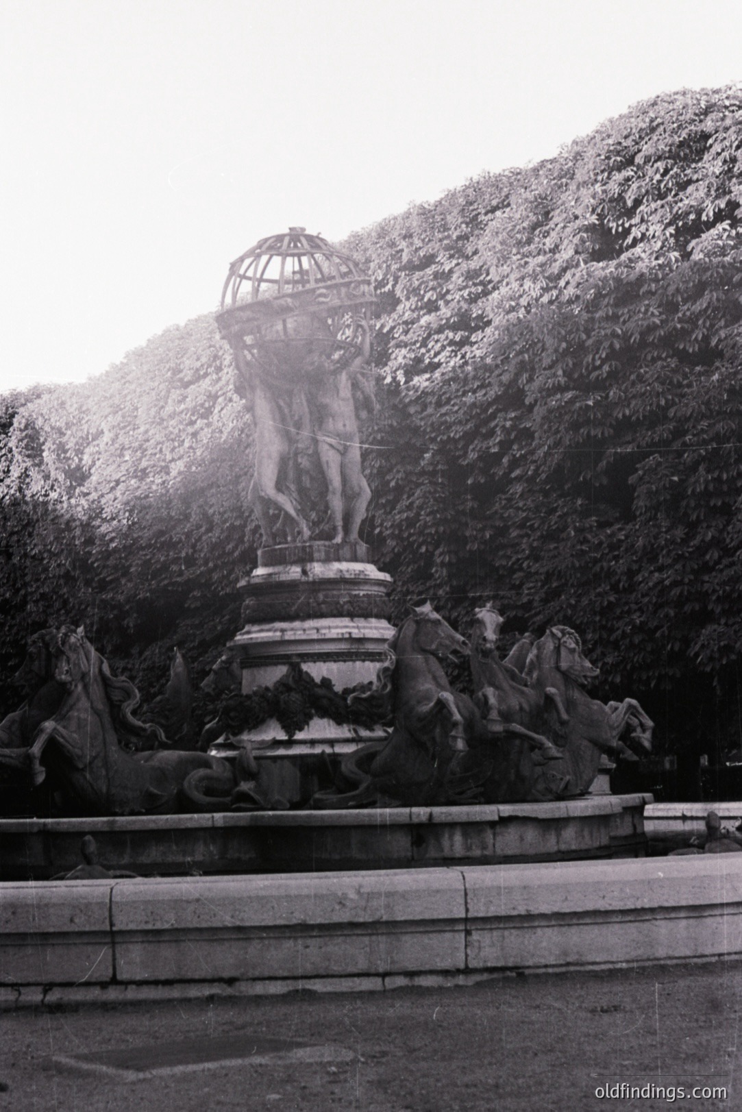 Monumental fountain depicts a muscular male figure holding aloft a tiered structure, surrounded by sculpted horses and water features. Likely 19th century, possibly European design influence. The fountain’s location, obscured by dense foliage, suggests a park or garden setting. The image shows signs of age with surface wear.