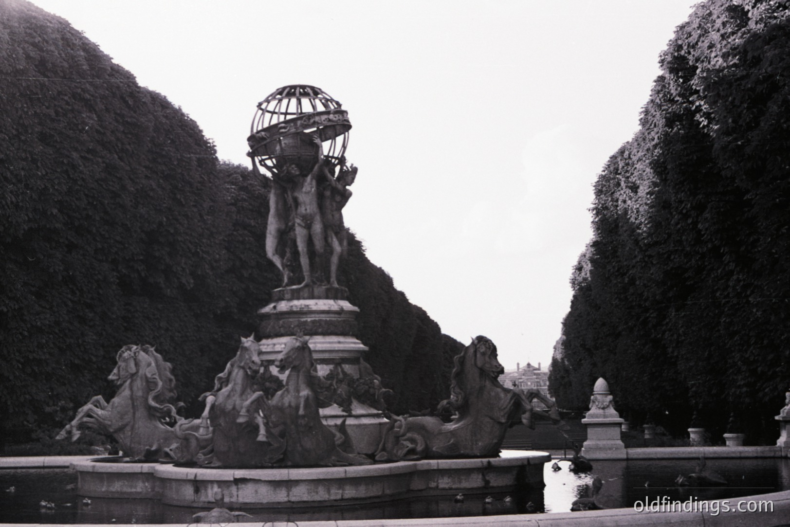 Ornate, tiered fountain features a sculpted male figure bearing a globe. Depicts sea creatures & horses on lower tiers within a formal garden setting, framed by sculpted hedges. Likely European, possibly 19th century. A stately, classical design for gardens or public spaces.