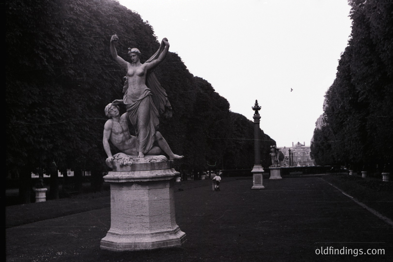 Dramatic neoclassical sculpture of a woman standing triumphantly over a fallen man, atop a stone pedestal. Formal, manicured hedges and a long perspective create depth. Likely a park or garden setting. Possibly 18th or 19th century in origin. Offers strong visual reference for historical design.