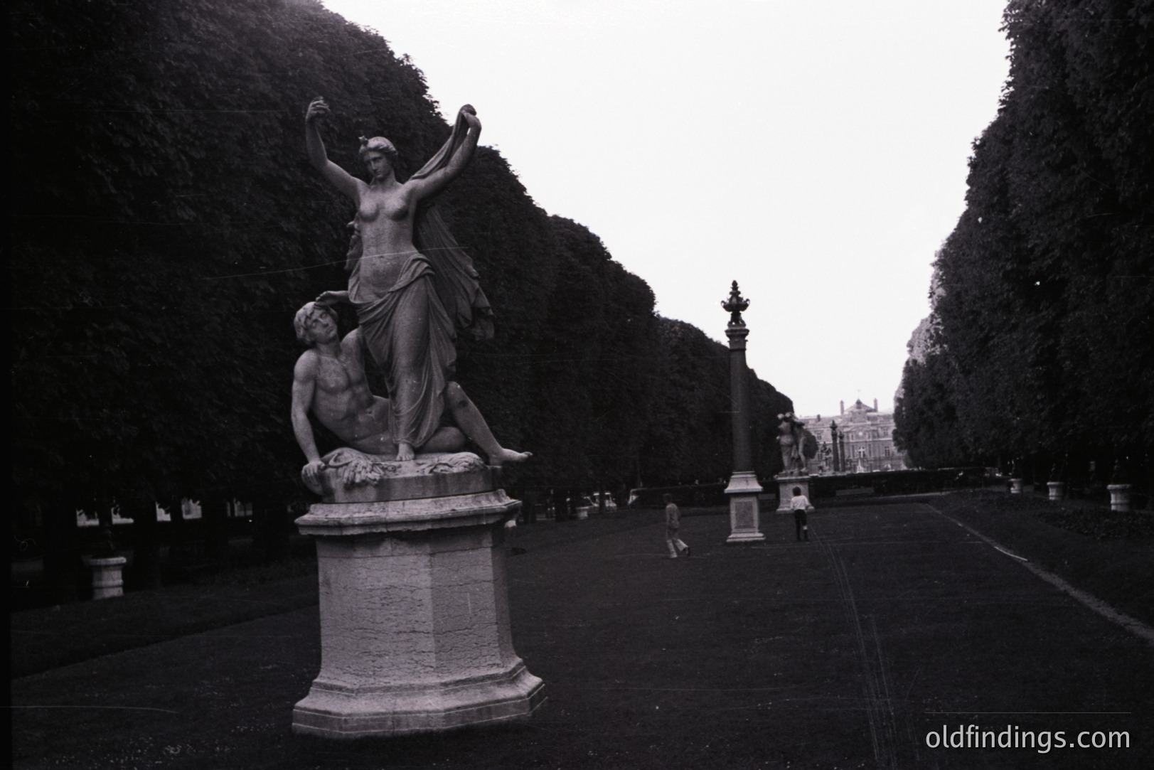 Ornate marble sculpture depicting a draped female figure supporting a fallen male figure. Formal, expansive park landscape with manicured hedges and distant architectural details. Likely a European estate or garden. Possible reference for classical art or landscape design.