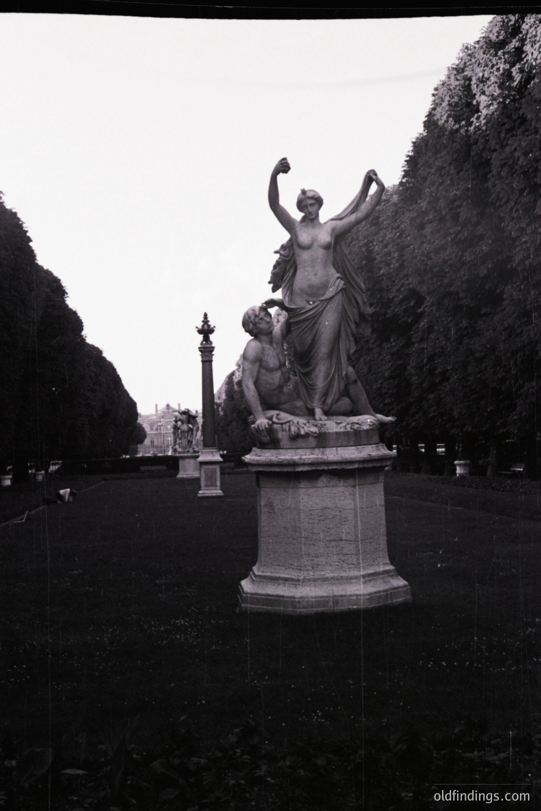 Classical sculpture depicting a robed female figure standing triumphantly over a fallen male figure. Likely depicting a mythological scene, set within a formal garden or park, featuring manicured lawns and dense trees. Possibly European origin, early 20th century based on photographic style.