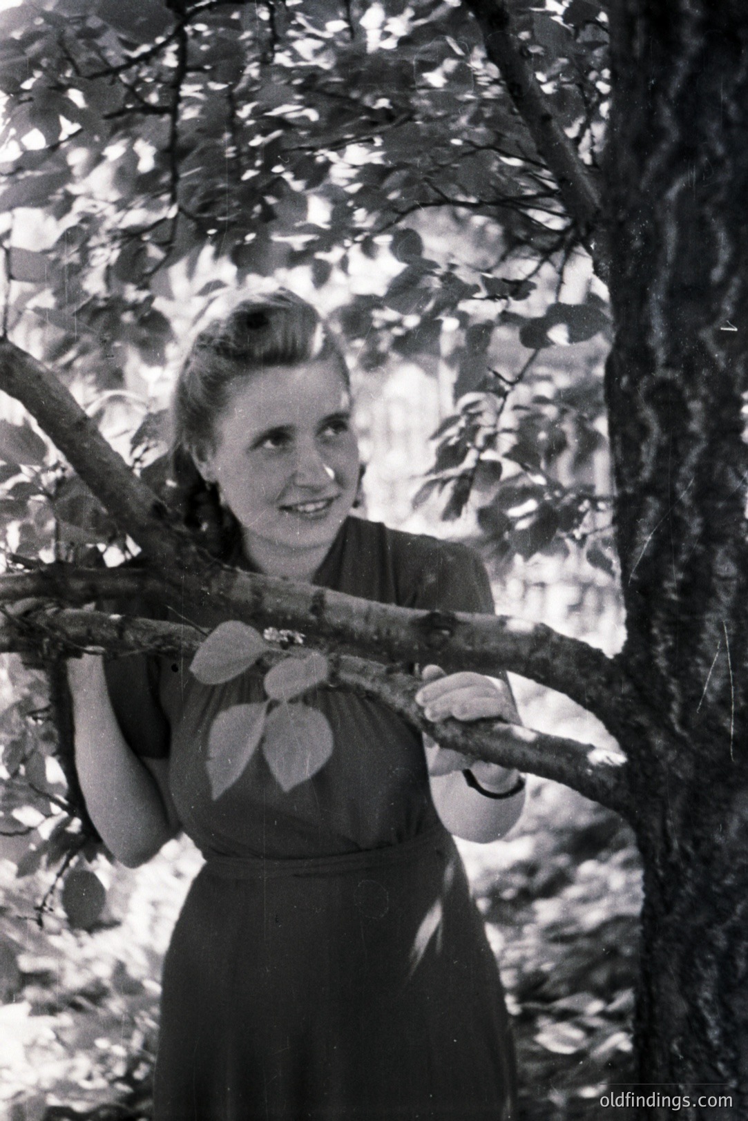 A young woman, seemingly in her late teens, smiles directly at the camera while leaning on a tree branch. She wears a modest, dark-colored dress with a fitted bodice and a gentle flare. The style suggests the 1940s or 1950s. Trees and foliage create a blurred backdrop. Likely a casual portrait.